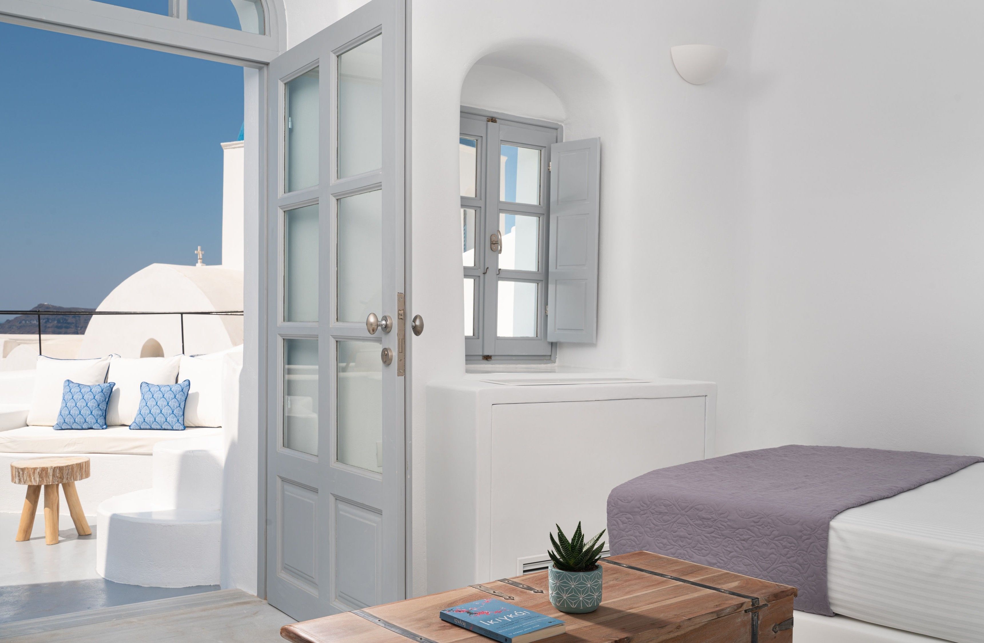 A bright, white room with a large window, a bed, a wooden table with a book and a plant, and a view of the sky and a building outside.