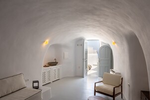Una habitación con paredes blancas y techo curvo, un sofá blanco, una silla de madera, una mesita pequeña y un armario blanco con un cuenco encima.