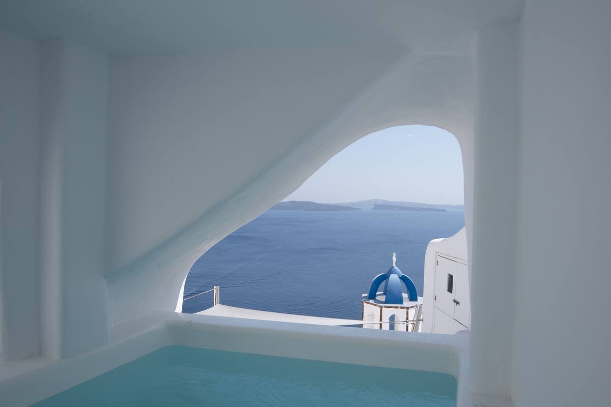 Blick von einem weiß getünchten Raum mit einem großen, bogenförmigen Fenster auf das Meer und ein blaues Kuppelgebäude.