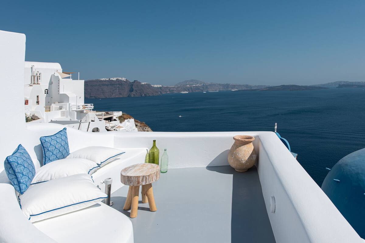 Ein weißer Balkon mit Meerblick, ausgestattet mit einer Couch, Kissen, einem kleinen Tisch und Dekoration.
