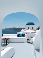 A white-walled room with a view of the sea and a blue-domed building.