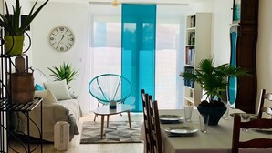 Una sala de estar con un sofá, una mesa de comedor para dos personas, una silla azul y un ventanal con cortina azul.