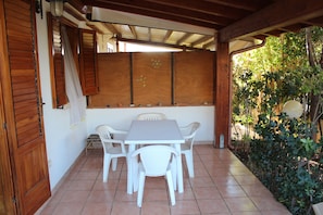 A covered patio with a white table and chairs, wooden shutters, and a view of greenery.