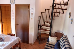 A room with a wooden door, a staircase with a black railing, a sofa with patterned cushions, and a table with a white tablecloth.