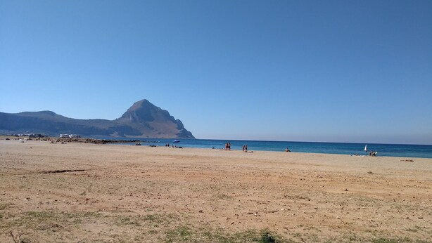 Una playa con una montaña al fondo, cielos despejados y algunas personas y vehículos.