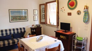 Un salón con un sofá a cuadros, una mesa de comedor con mantel blanco, un mueble de madera, un televisor y una estantería empotrada con objetos decorativos.