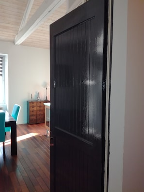 Una habitación con piso de madera, una puerta negra, una mesa de comedor con sillas y un mueble con objetos decorativos.