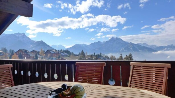 A wooden deck with a table and chairs, overlooking a mountainous landscape with houses and a clear sky. - Sunset B 11 by Interhome (Villars)