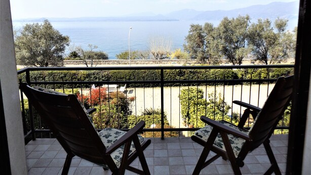 Un balcón con dos sillas de madera, piso de baldosas y vista al mar y a las montañas.