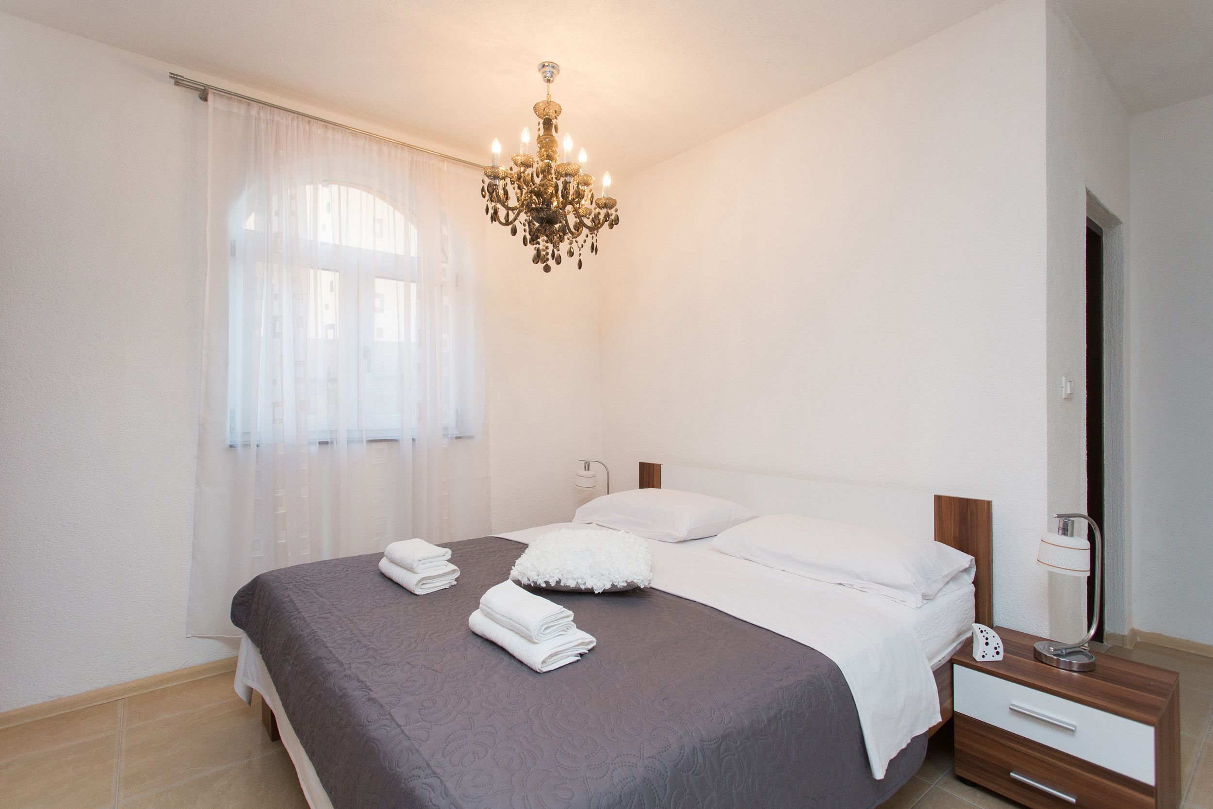 A neatly made bed with white linens, a chandelier, and a bedside table with a lamp.