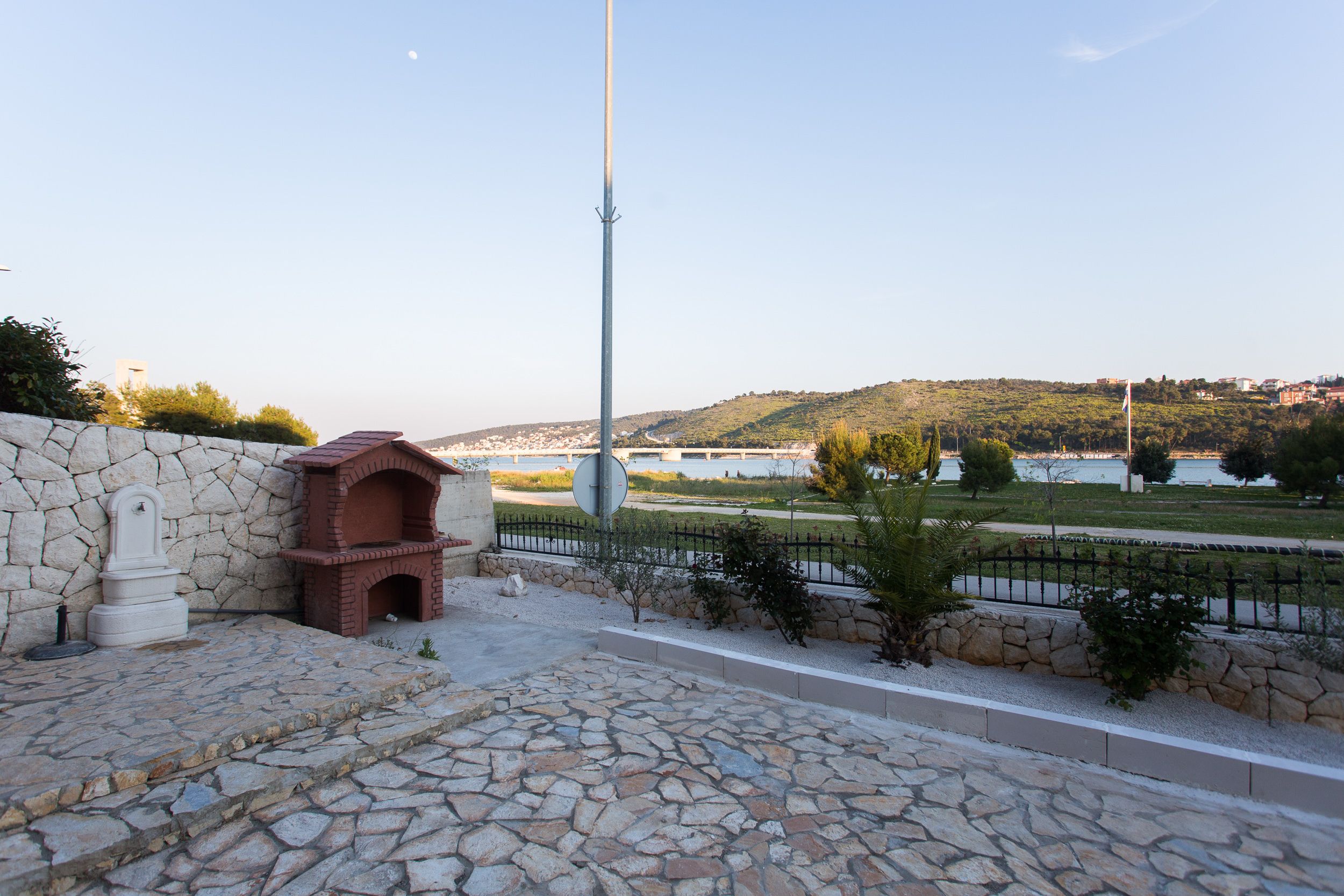 A stone pathway leading to a brick structure, a metal pole, and a view of a river and hills in the background.
