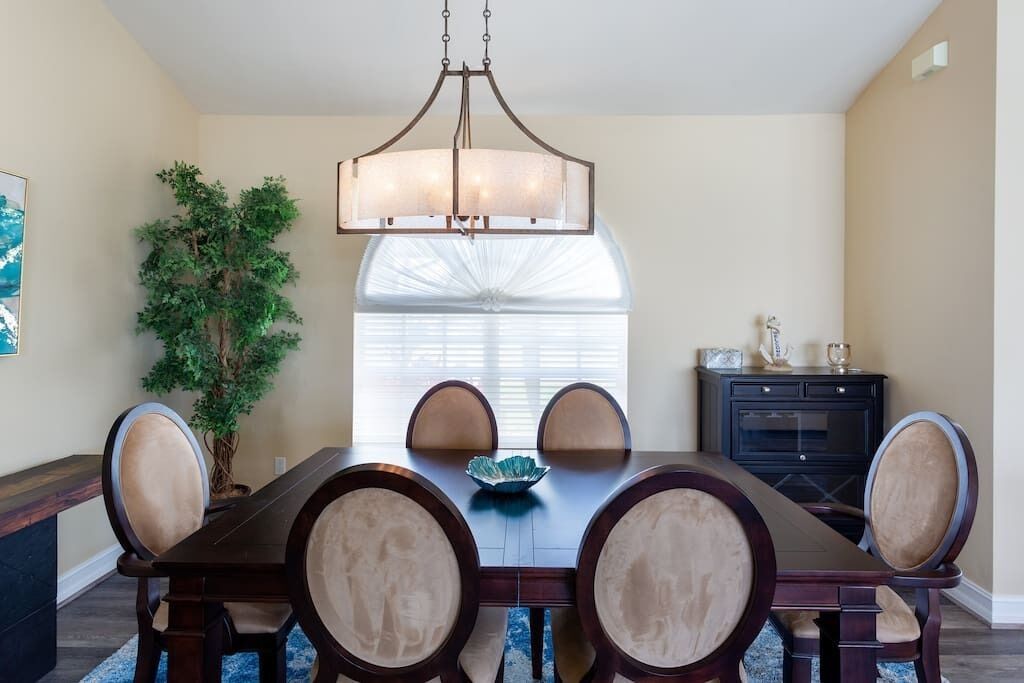 Une salle à manger avec une table en bois foncé, des chaises recouvertes d’un tissu beige, un lustre, une plante en pot et une fenêtre avec des stores.