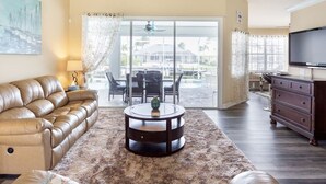 Un salon avec un canapé en cuir, une table basse ronde, un téléviseur et une vue sur une terrasse extérieure, accessible par une porte vitrée.