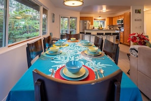 A dining area with a table set for six, surrounded by chairs, with a view of the kitchen and a dining table with a blue tablecloth.