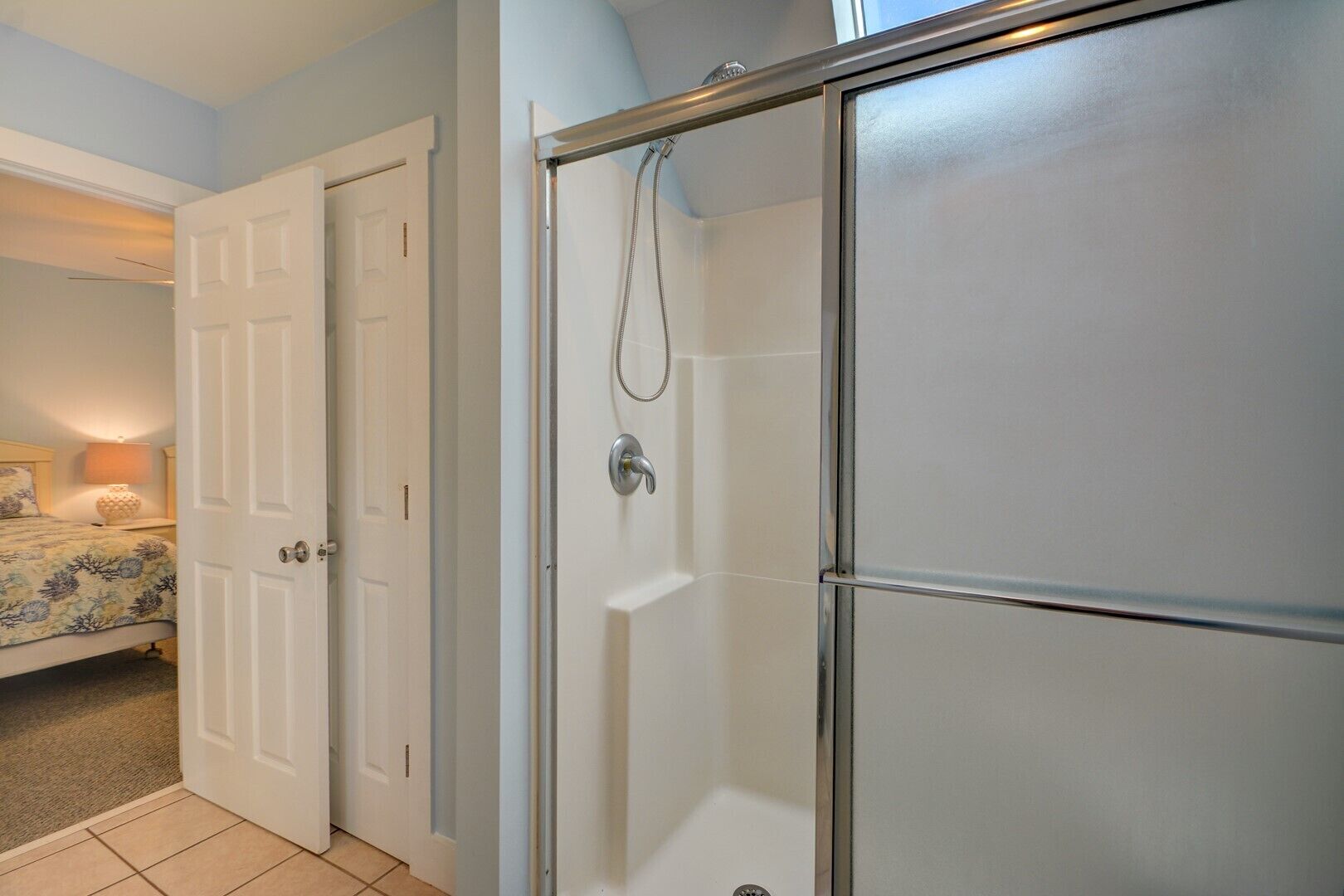 Un baño con cabina de ducha de vidrio, una puerta blanca y una cama con edredón estampado floral.