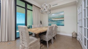 A dining area with a wooden table and wicker chairs, a chandelier, a painting of a boat, and a glass door with a view. - Gulf Glow & Lazy River Flow - Phoenix West 1102 (Orange Beach)