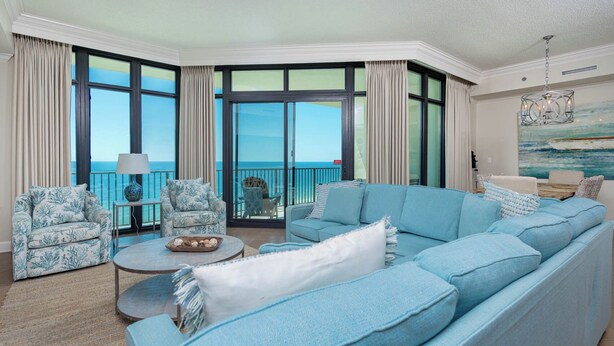 A spacious living room with a large blue sofa, two patterned armchairs, a round coffee table, and a view of the ocean through large windows. - Gulf Glow & Lazy River Flow - Phoenix West 1102 (Orange Beach)