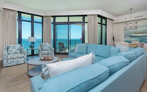 Un salón espacioso con un amplio sofá azul, dos sillones estampados, una mesa de centro redonda y vistas al océano a través de grandes ventanales.