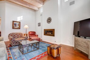 A living room with a patterned sofa, a glass coffee table, a leather armchair, a television, and a fireplace.