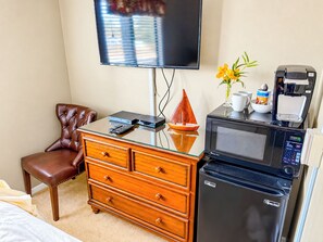 Quarto de hotel com TV de tela plana na parede, cómoda de madeira com gavetas, cadeira de couro marrom, micro-ondas e máquina de café no balcão.