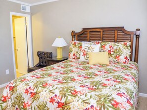 Quarto com colcha florida, cabeceira de madeira, mesa de cabeceira com abajur e poltrona de couro.