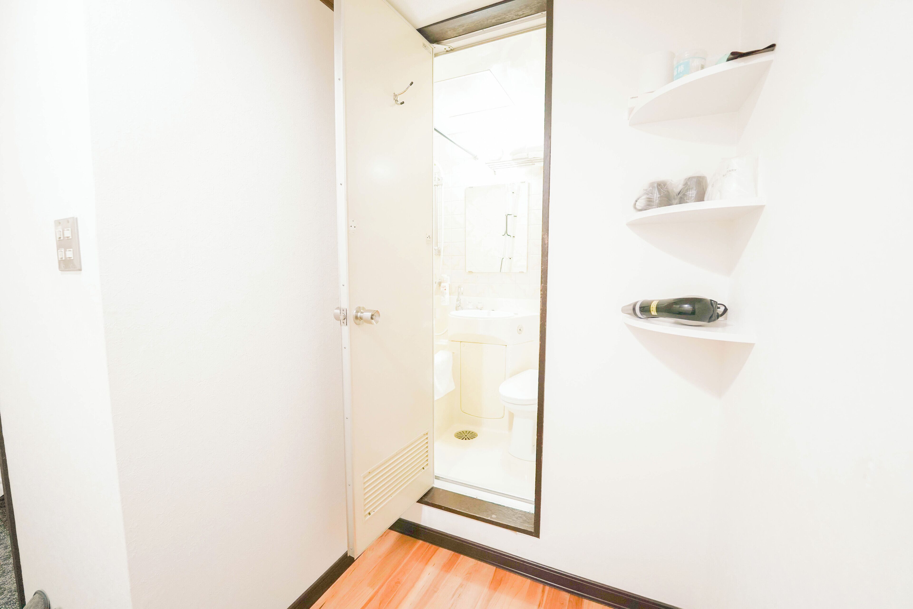 A hallway with a door leading to a bathroom, a wooden floor, and white walls with a shelf holding toiletries.
