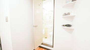 A hallway with a door leading to a bathroom, a wooden floor, and white walls with a shelf holding toiletries.