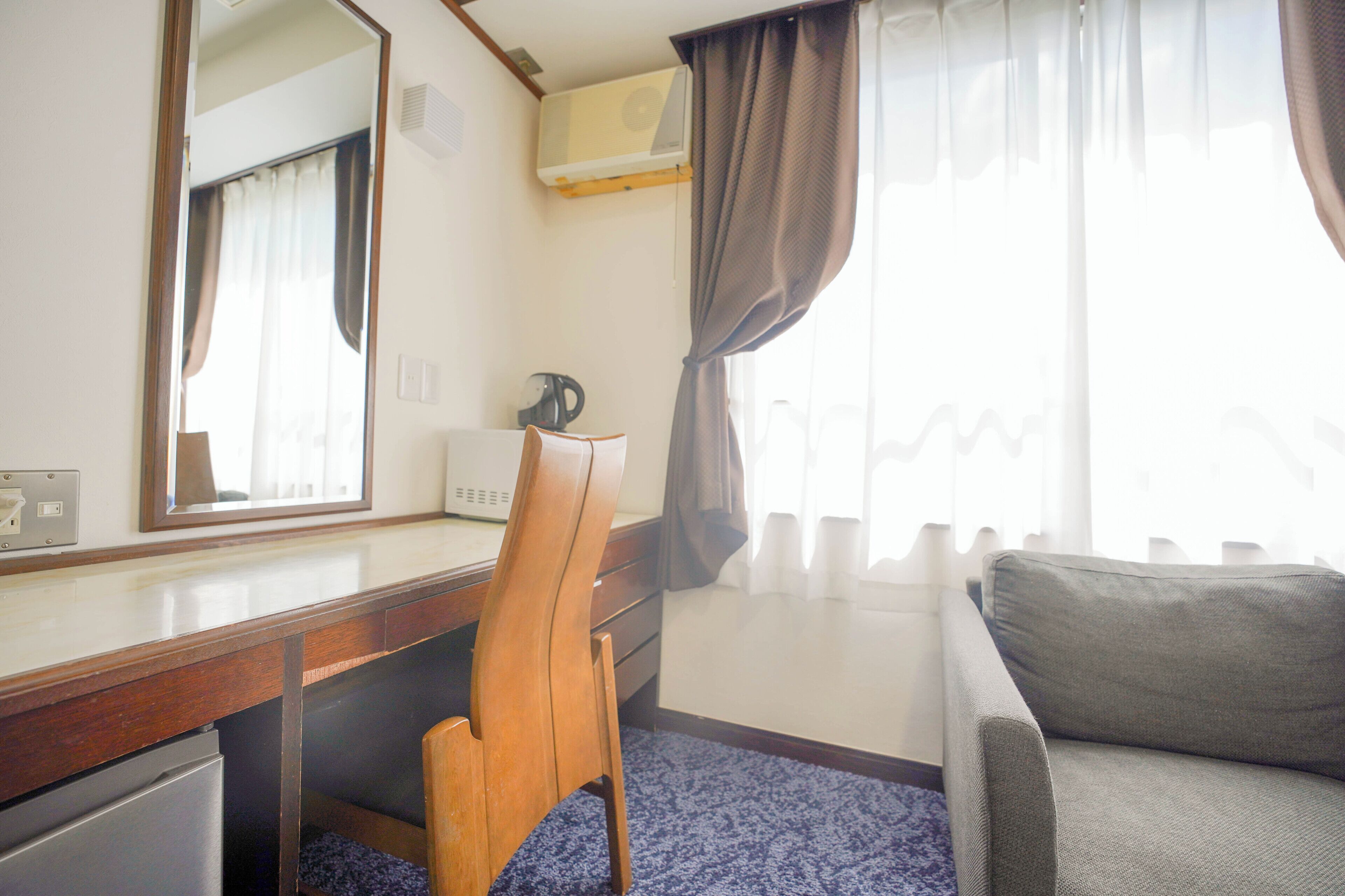 A hotel room with a desk, chair, sofa, air conditioning unit, and a window with curtains.