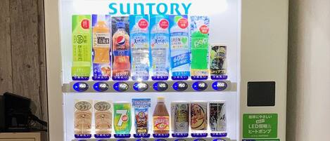 Máquina expendedora con diversas bebidas y aperitivos, incluyendo marcas como Pepsi y 7UP, y un letrero verde que indica servicio las 24 horas.