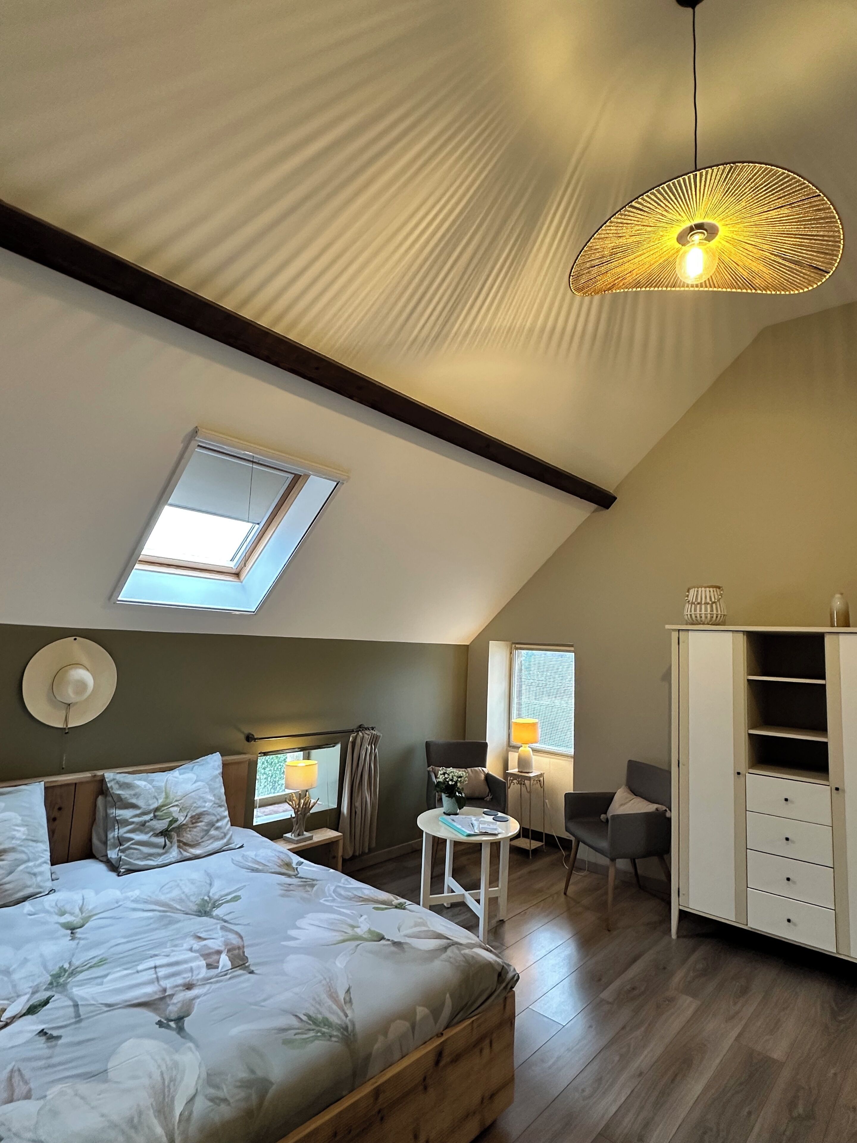 Un dormitorio con techo inclinado, tragaluces, cama con ropa de cama floral, mesita de noche, silla, escritorio, lámpara, armario y una lámpara colgante.