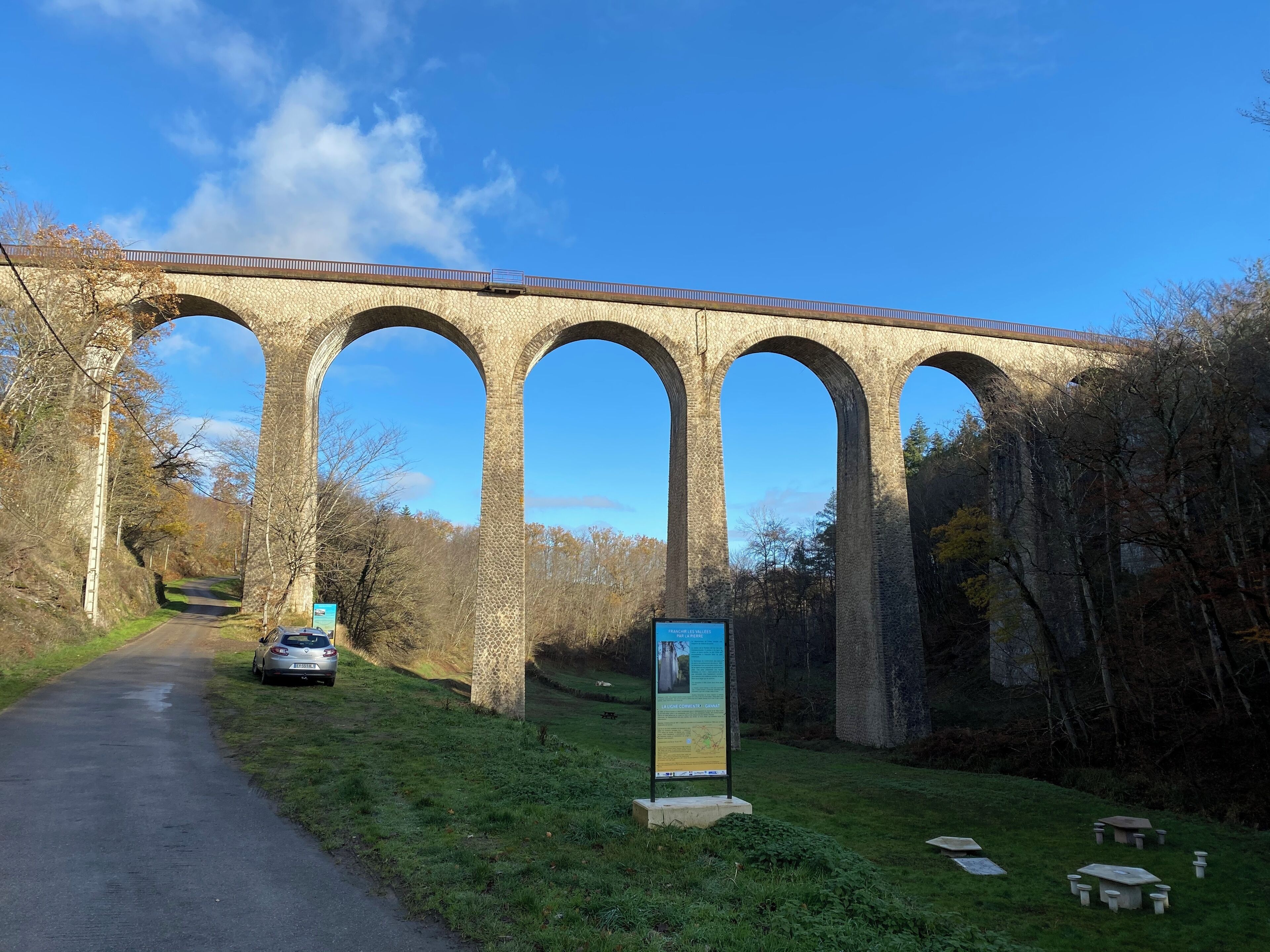 Un viaducto de piedra con múltiples arcos sobre una carretera, rodeado de árboles y una zona verde con mesas de picnic.