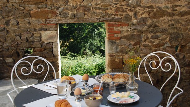 A round table with breakfast items, including croissants, eggs, and bread, set against a stone wall with a view of greenery. - B&B Maison Balady (Bellenaves)