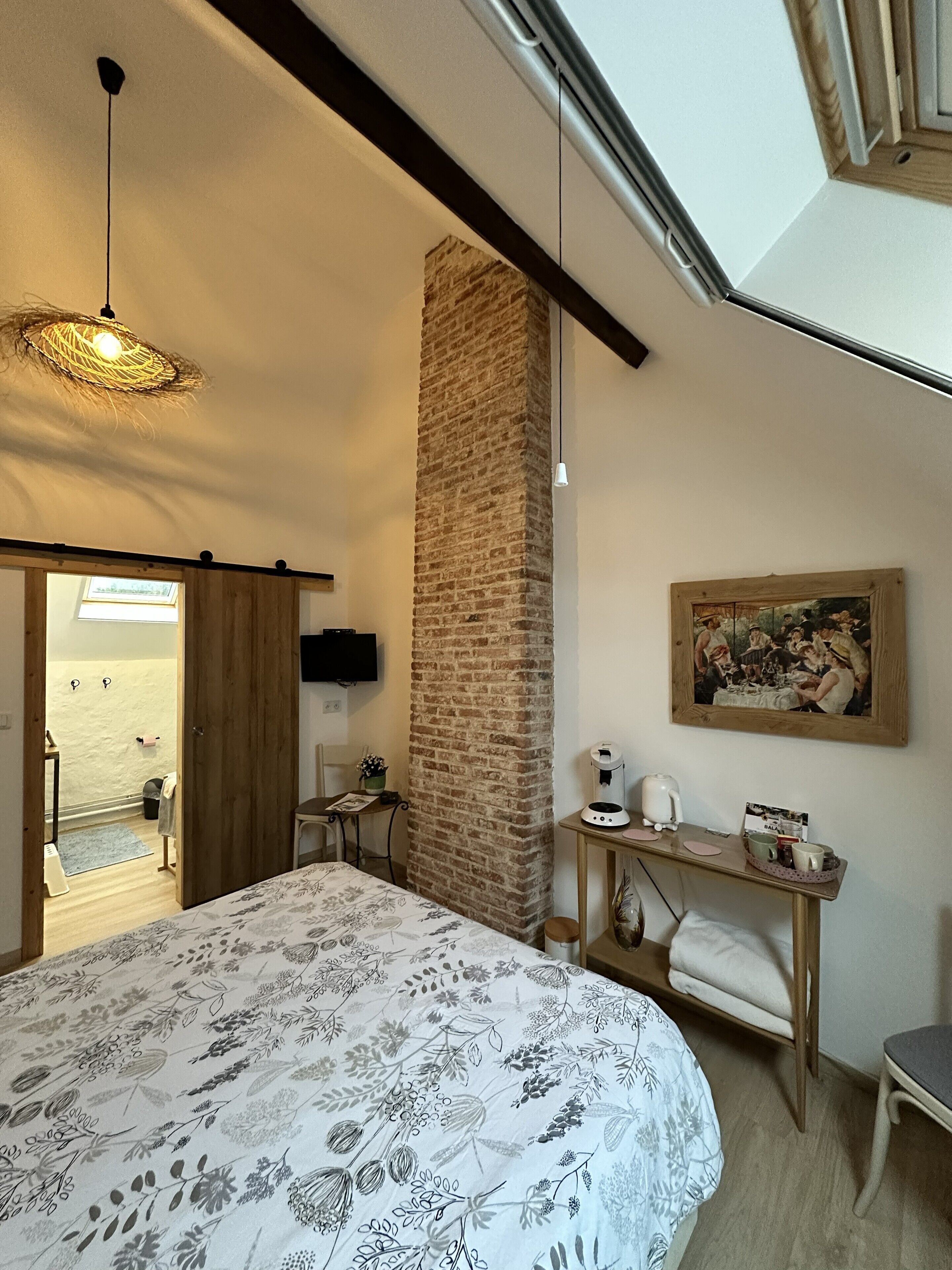 Une chambre avec un grand lit, un pilier en pierre, un bureau avec une machine à café, un tableau et une lucarne.