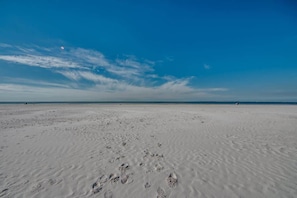 A wide sandy beach with footprints, a clear blue sky, and a calm sea.
