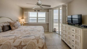 Un dormitorio con cama, televisor, ventilador de techo, ventana con contraventanas y vista al océano.