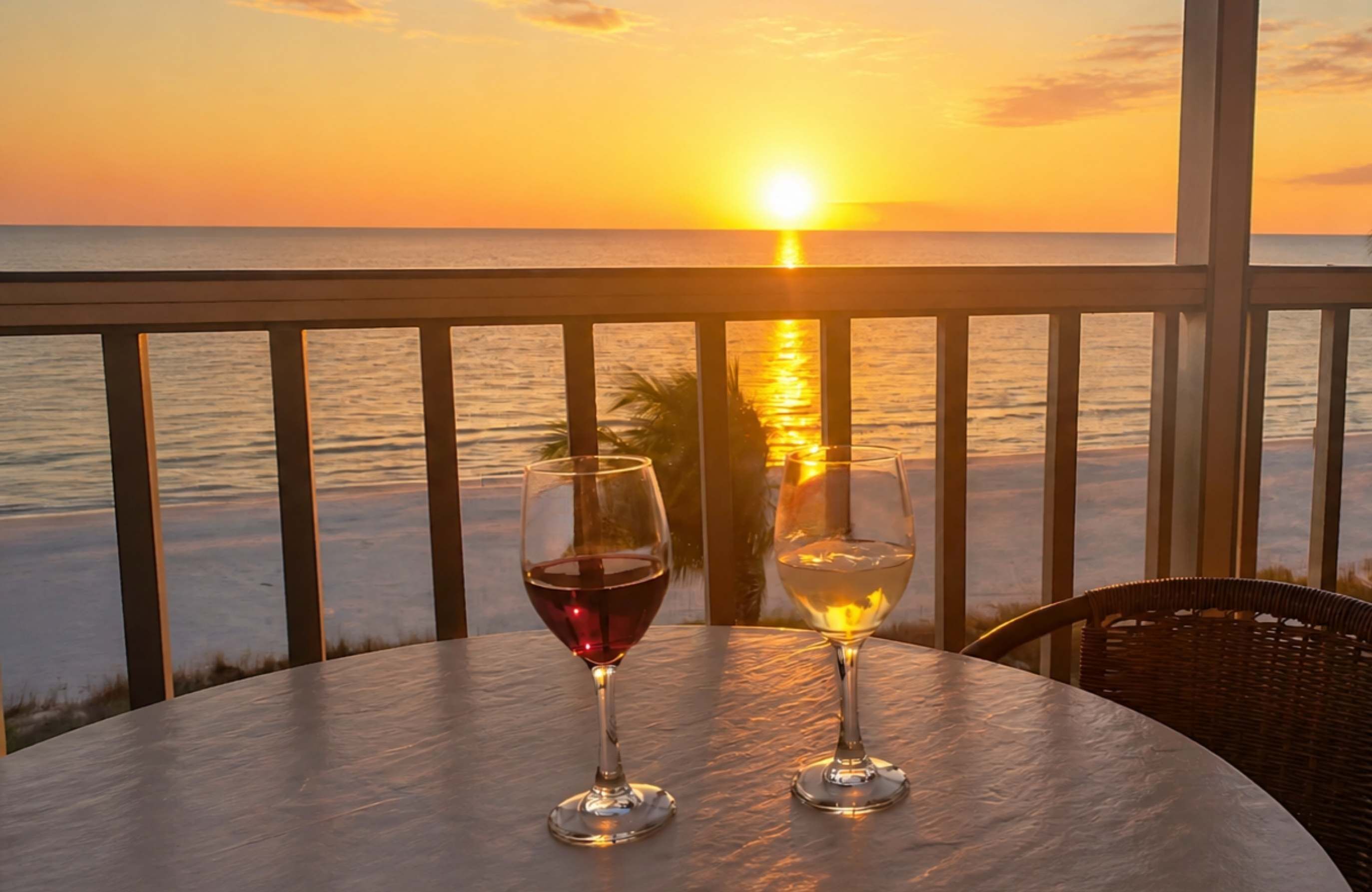 A table with two wine glasses on a balcony overlooking a sunset over the ocean.