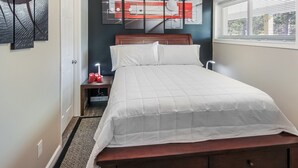 Ein Schlafzimmer mit Bett, Nachttisch, Wandbild und einem Fenster mit Jalousien.