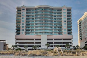 A multi-story hotel with a curved facade, multiple balconies, and a parking garage below. - Beautifully remodeled, oceanfront condo + Free Attraction Tickets! (North Myrtle Beach)