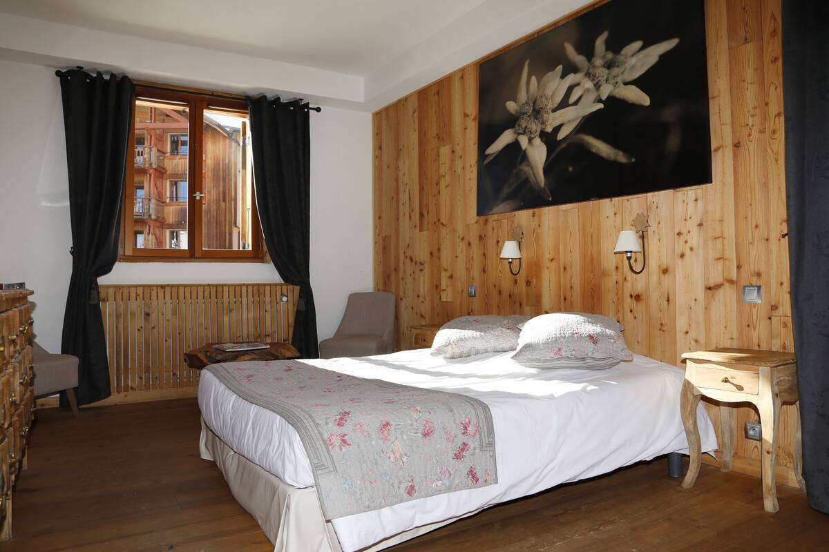 A bedroom with a wooden paneled wall, a bed with a floral bedspread, a window with curtains, and a wooden dresser.