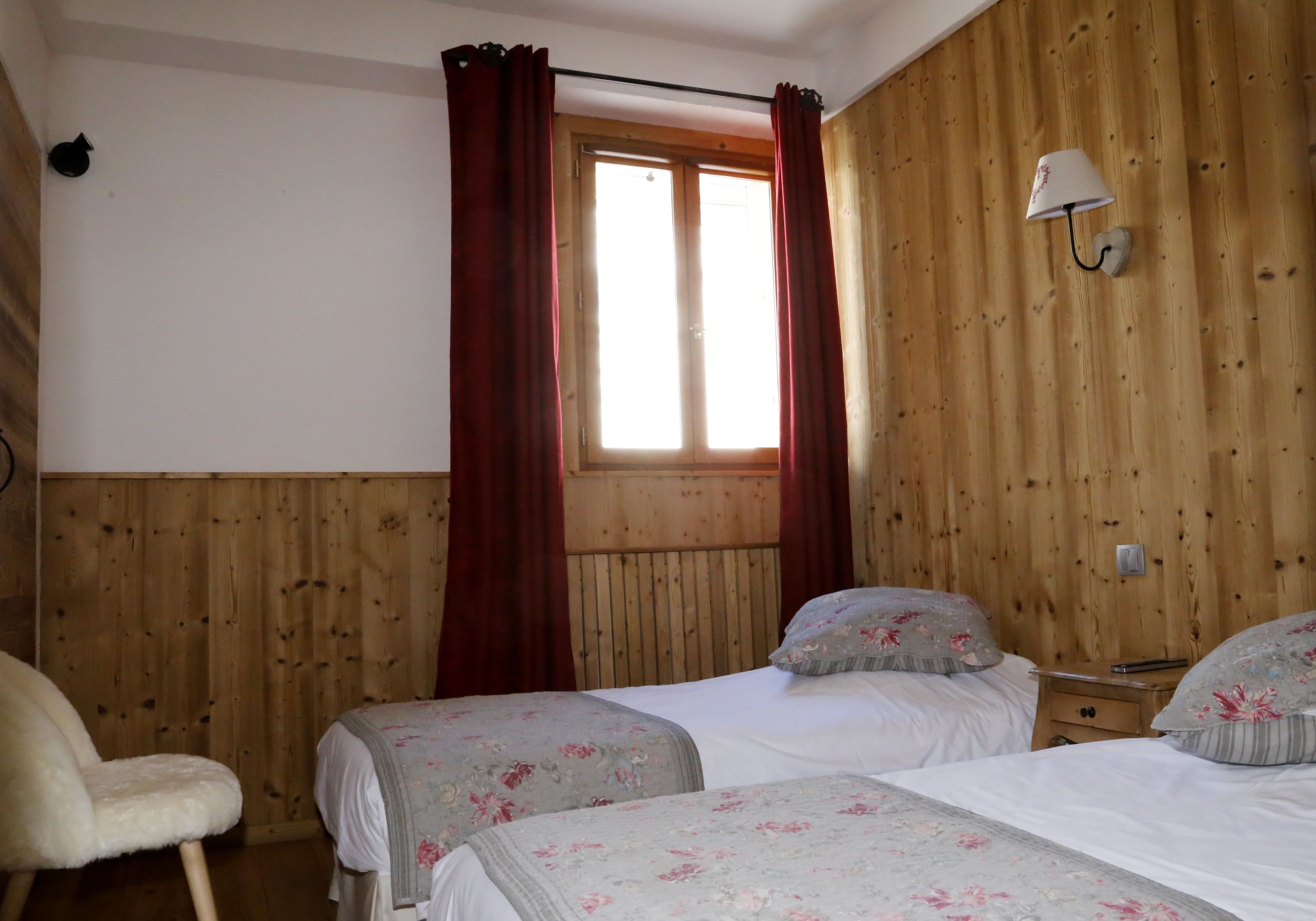 A bedroom with two beds, wooden walls, a window with red curtains, and a nightstand with a lamp.