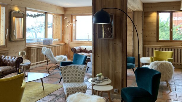 A spacious room with wooden walls, a large window, and a variety of seating including a leather couch, patterned chairs, and a fluffy stool. - Alp'Azur Hotel (Huez)