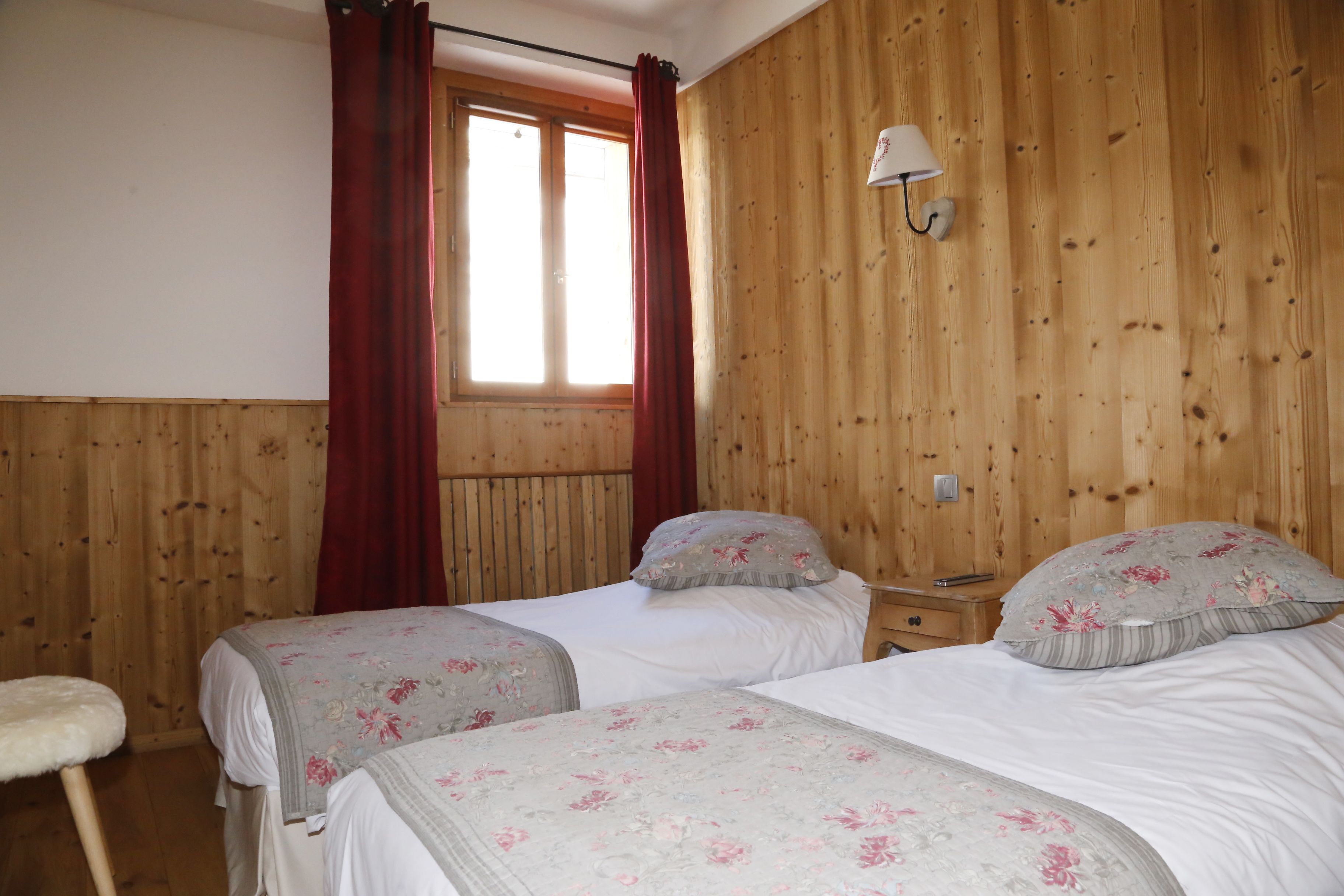 A room with two beds, wooden walls, a window with curtains, a nightstand, and a wall-mounted lamp.