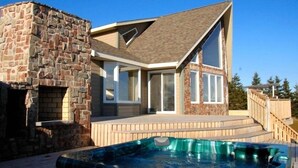 A modern house with a stone fireplace, a large deck, and a hot tub. - Luxury House in the Cavendish Area with Pool, Hot Tub and Air Conditioning (Stanley Bridge)