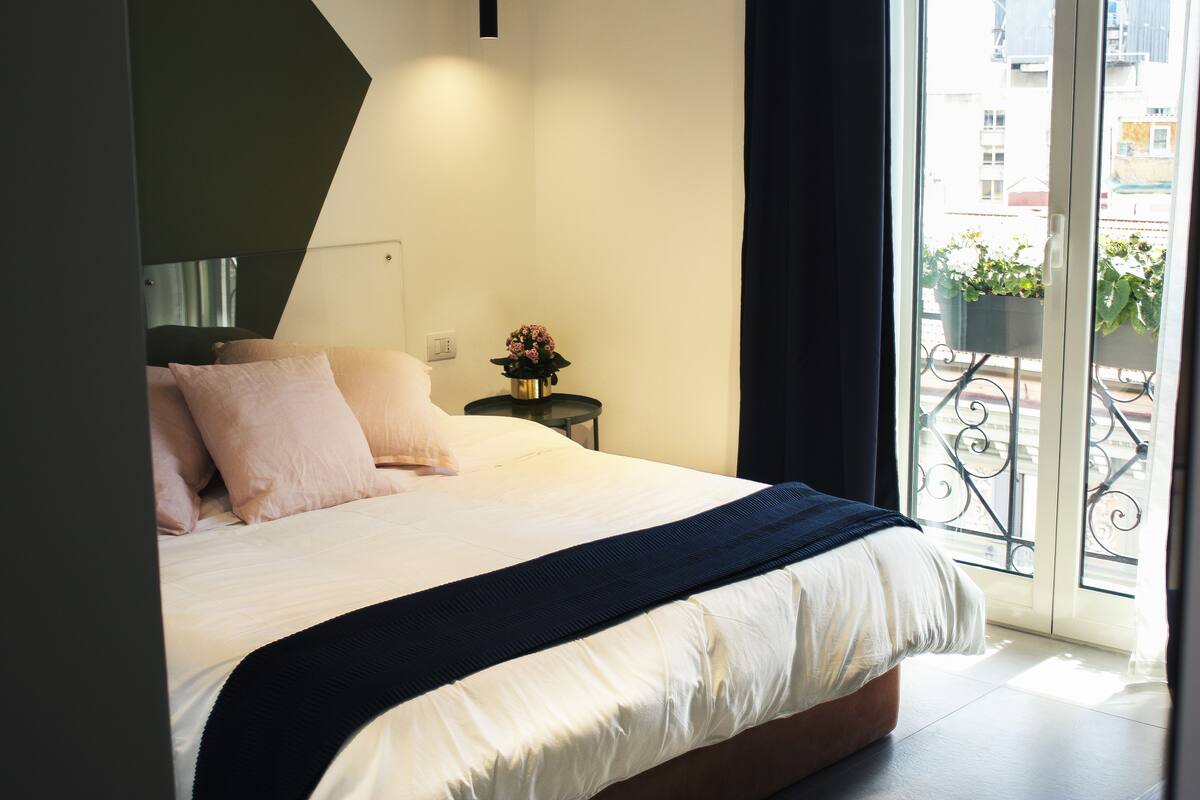 Une chambre avec un lit, une table de chevet, une plante en pot et une vue sur le balcon extérieur.