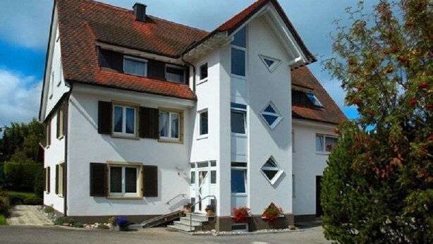 Une maison à deux étages, avec un toit recouvert de tuiles rouges, des murs blancs et un petit porche à l’avant, accessible par quelques marches.