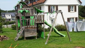Una casita de juegos de madera con tobogán y zona de escalada, ubicada en un patio cubierto de césped con una casa blanca al fondo.