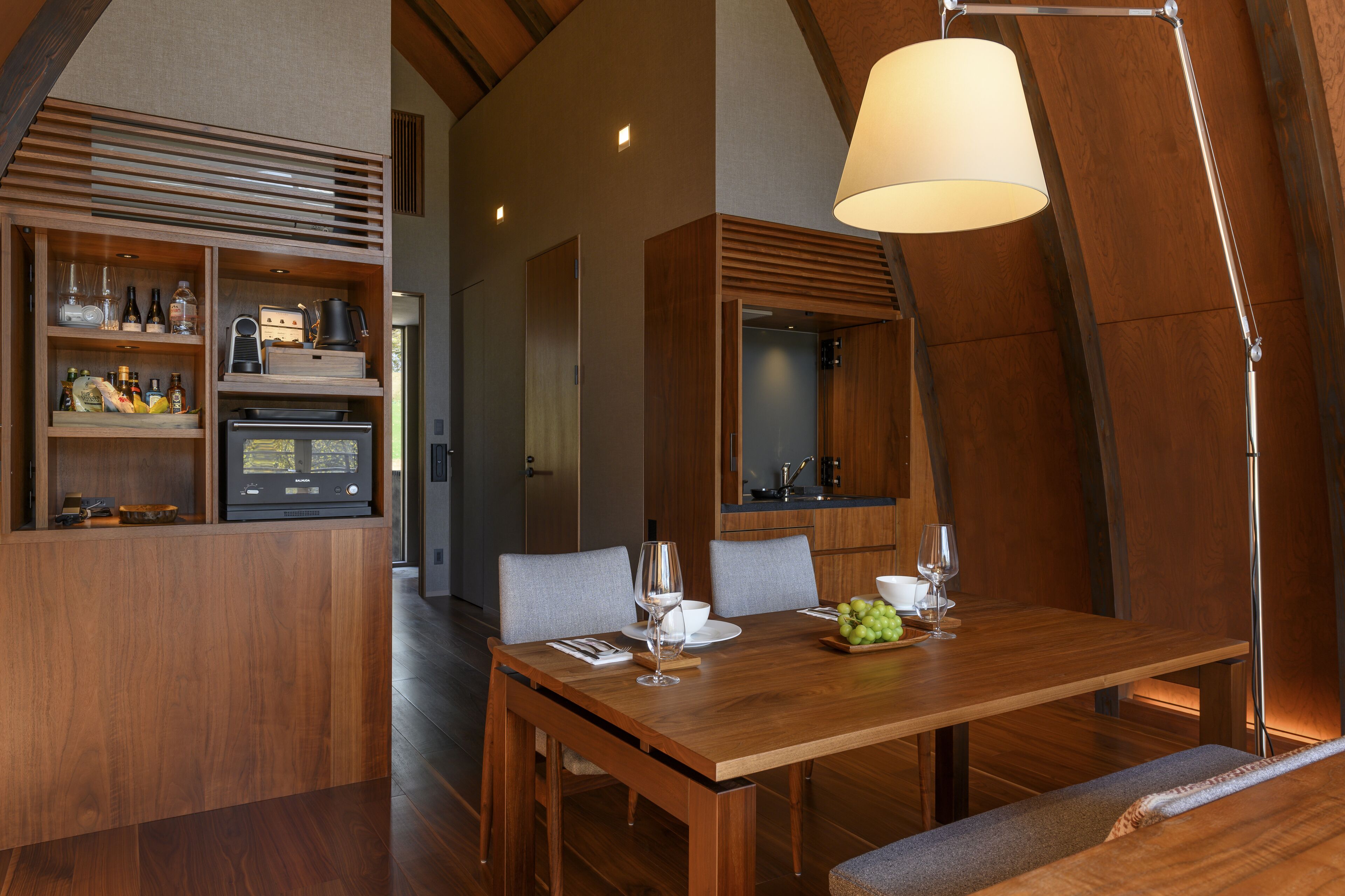 Een moderne eethoek met een houten tafel, stoelen en een lamp. Er is een keuken met kastjes en een magnetron.