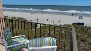 A balcony with two striped chairs overlooking a beach with people and umbrellas. - 2 night minimum. Discounts all year. 1 bedroom oceanfront. Great location. Pool. (North Myrtle Beach)