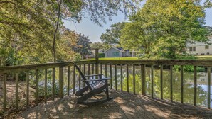A wooden deck with a rocking chair overlooking a calm lake, surrounded by lush trees and a house in the background. - Charleston-area 3BR, Close to Beach or Downtown, Walk to Shem Creek (Mt Pleasant)