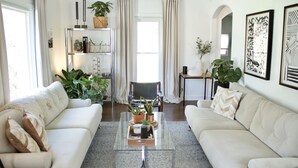 A living room with two white sofas, a glass coffee table, and a bookshelf with decorative items. - Walk to Journeyman Renovated Spacious Victorian House, Beautiful Vintage Details (Three Oaks)